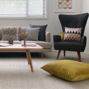 A room styled in mid century modern decor with a beige sofa and large charcoal chair. There are cushions in yellow and brown colours and a mustard floor cushion which gives a retro and vintage vibe.