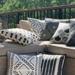 Black and white outdoor cushions are stacked and styled on an outdoor corner chair in front of a vibrant green bush backdrop. The tribal design of the covers share dots, strokes and decorative patterns.