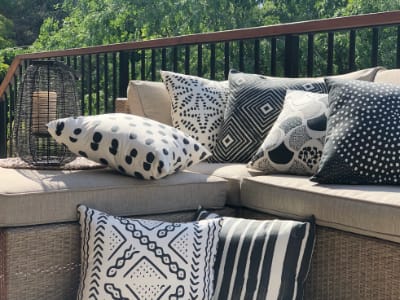 Black and white outdoor cushions are stacked and styled on an outdoor corner chair in front of a vibrant green bush backdrop. The tribal design of the covers share dots, strokes and decorative patterns.