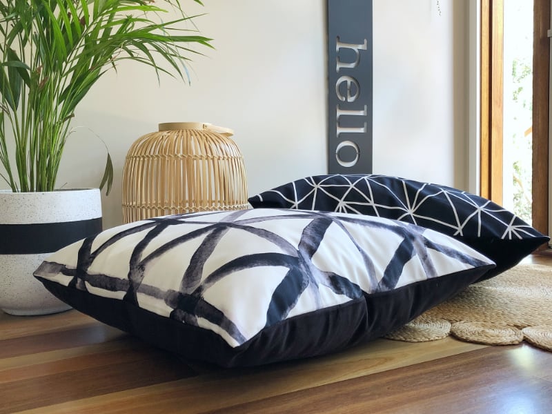 Two black and white velvet floor cushions are styled on a wooden floor with a green fern in the background