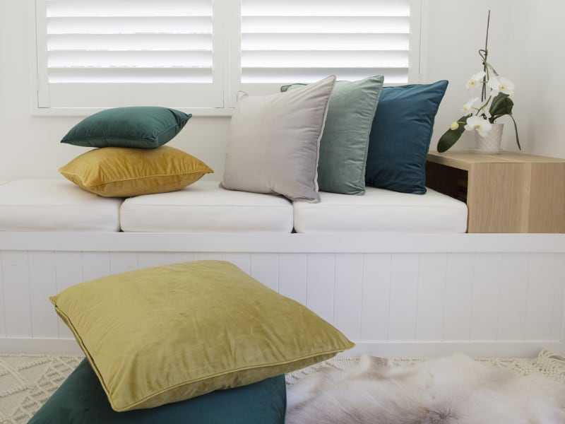 A white room is shown with large cushions layered on the floor and the nearby day bed which sits under a window