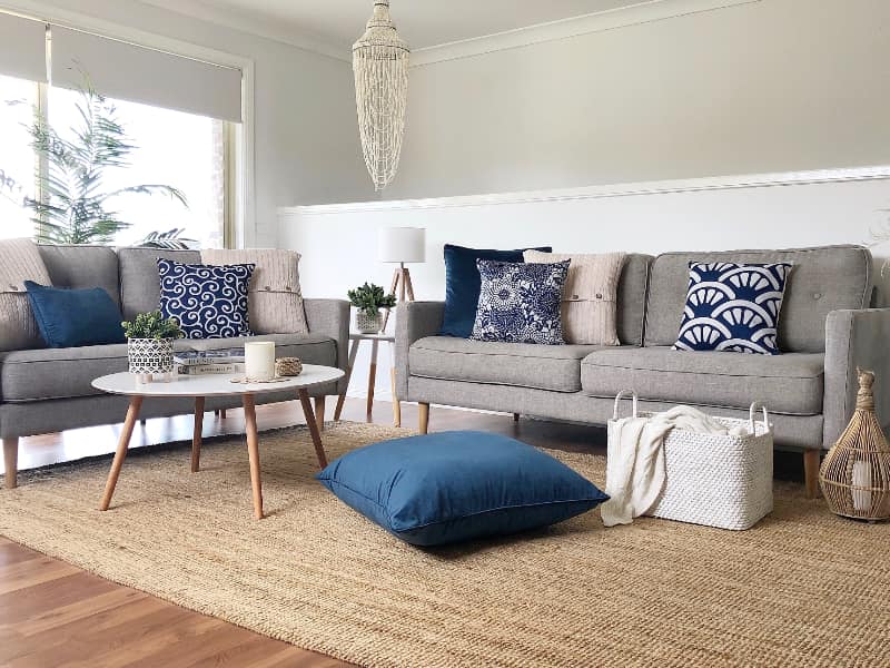 A vivid coastal scene is shown with two large grey sofas displaying a series of blue cushion covers styled across them and on the floor