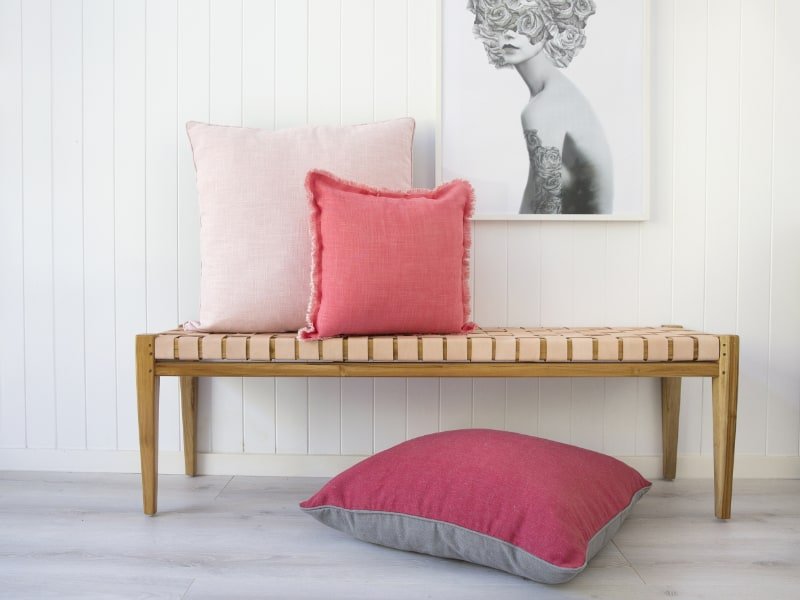 A collection of pink cushions is shown in a light coloured scene with artwork hanging gin the background