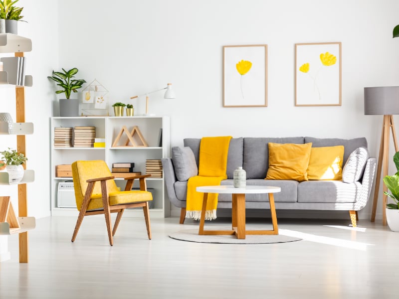 A selection of yellow cushion covers is shown arranged on a grey sofa in a bright and airy white coloured room with yellow featuring also in the nearby throw, arm chair and on the artwork on the wall