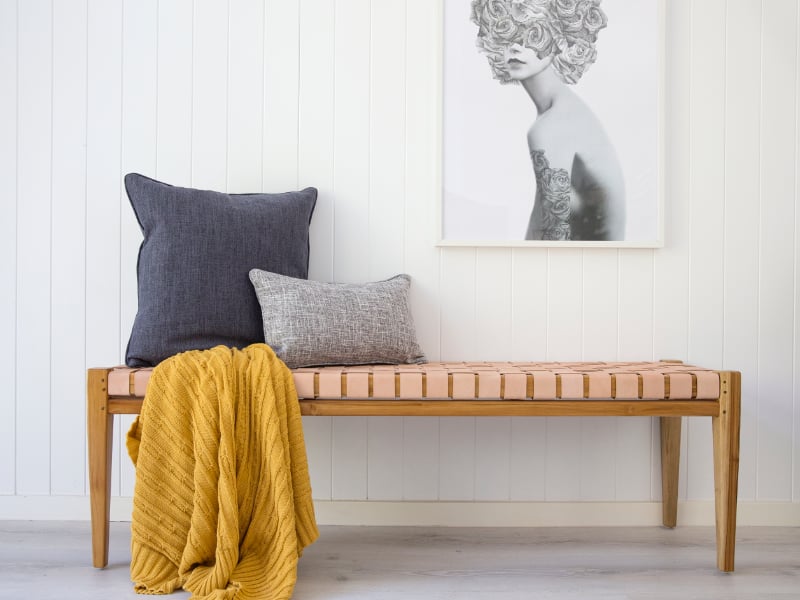 A pair of grey cushions sit on a beautiful wooden bench and are partnered with a lavish mustard throw blanket while a monochrome artwork sits behind on a wall