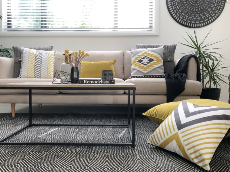 A light coloured sofa is shown with grey and yellow cushions placed across it and a dark charcoal and white rug sits underneath a modern coffee table and stack of floor pillows