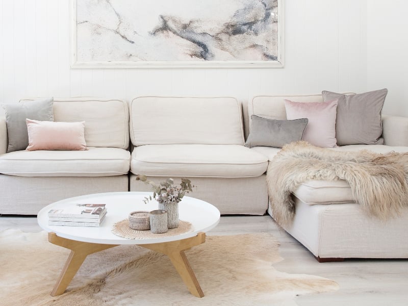 A bright modern scene features pink and grey cushions on a light coloured couch with various furs and hides nearby and an enchanting piece of artwork is displayed above the sofa
