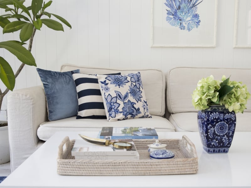 In this picture an elegant couch is shown that has several Hamptons style cushions placed on it and a white coffee table in to the front of it with a small potted plant and numerous books in a serving tray