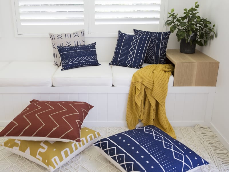A white room is pictured with a white window seat dressed with bright colourful cushions arranged on it and on the floor