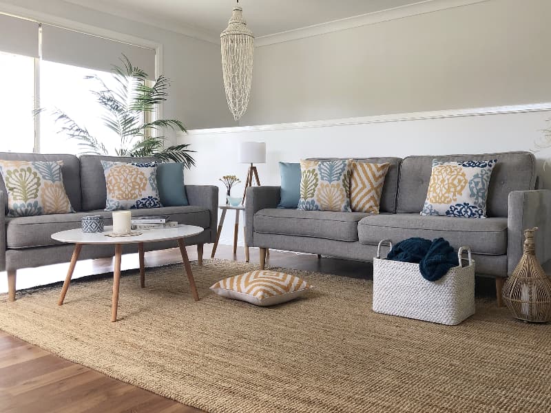 A beautiful lounge room from a holiday beach house is shown with two grey sofas hosting a delightful collection of coastal cushions in teal yellow and sage green colours