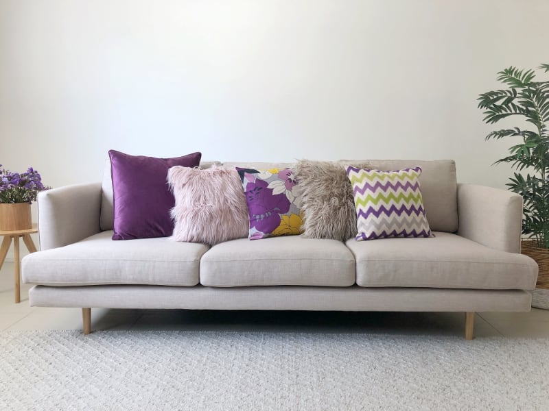 A selection of decorative cushions in purple and soft pinks tastefully arranged on a grey sofa with potted indoor plants to the side