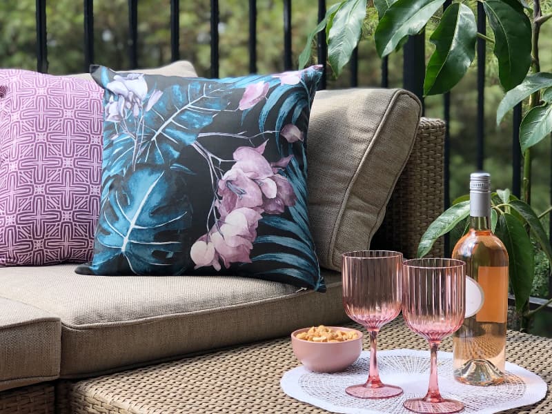 In this image an outdoor sofa is shown decorated with an arrangement of floral cushions in pink and blue while a small table sits in the foreground with a bottle of wine and glasses
