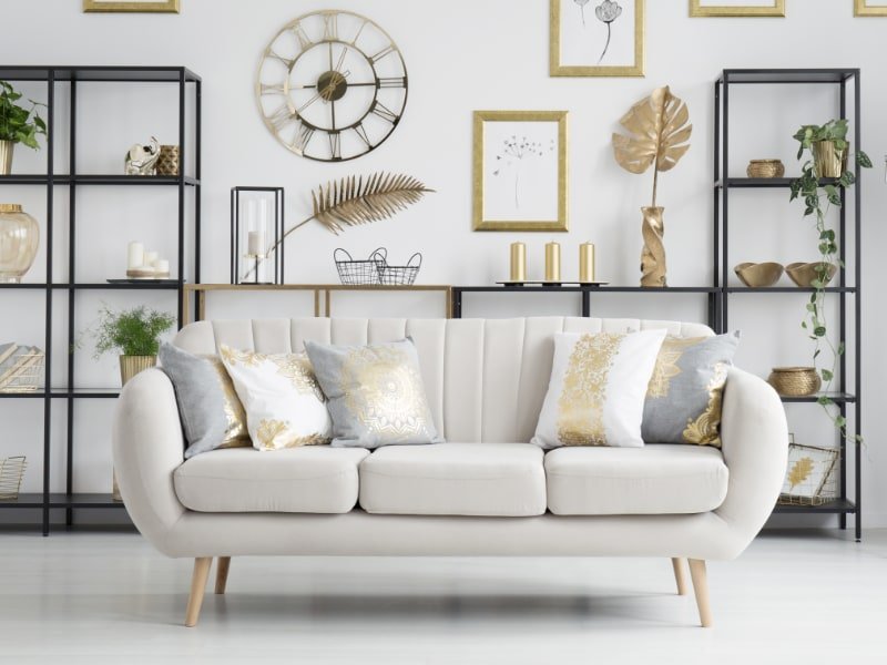 A stylish contemporary room is shown with a light grey sofa that features a collection of gold cushion covers surrounded by black metal framed shelving, numerous gold decor items and wall hangings