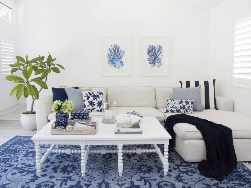 A bright and airy sitting room is shown with sea-inspired decor, white furniture and a light coloured sofa that has a collection of blue and white Hamptons cushions arranged upon it with a nearby dark navy throw blanket
