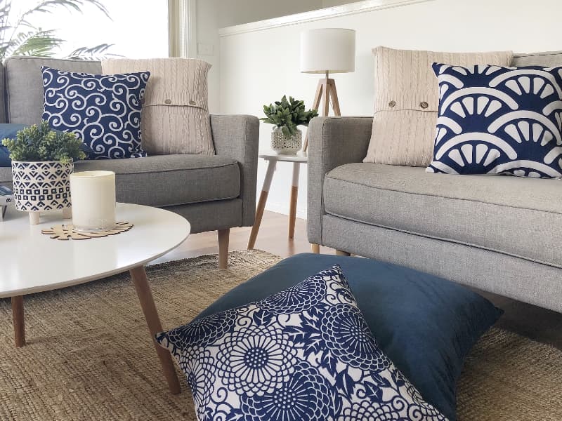 A bright and airy beach house is pictures with an elegant display of white and navy cushions arranged on a light grey sofa with other white decor items and furniture adding to the coastal look
