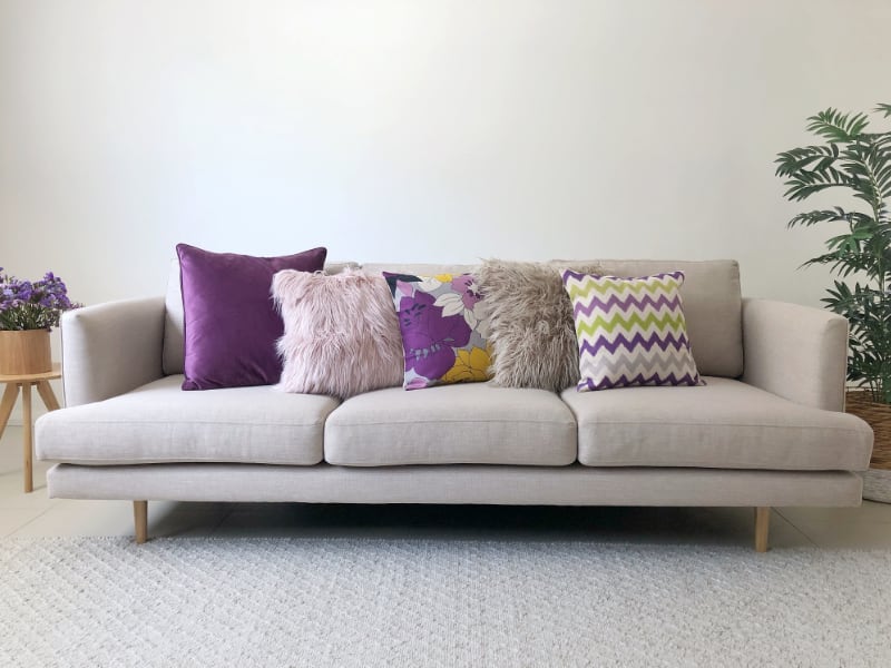 A bright room is pictured with a 3 seat sofa centred with a collection of covers in various textures including a pink fluffy cushion nestled amongst vibrant purple styles