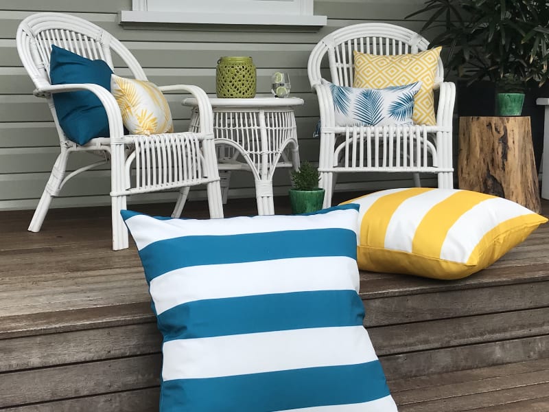 A wooden backyard patio scene is pictured showing a collection of elegant and vibrant yellow and teal outdoor cushions arranged in and around some white furniture