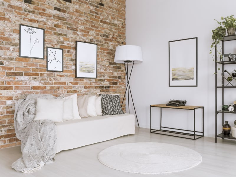 An urban industrial themed room is shown with a collection of white cushion covers displayed on a light coloured seat with an exposed brick backdrop and ornaments featuring metal hanging on the walls nearby