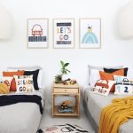 Kids cushions wit cute car designs on them in blues and reds sitting on twin beds with a mustard and navy throw blanket. On the wall are three kids wall prints that match the designs on the cushions