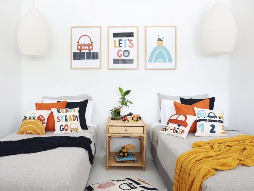 Kids cushions wit cute car designs on them in blues and reds sitting on twin beds with a mustard and navy throw blanket. On the wall are three kids wall prints that match the designs on the cushions
