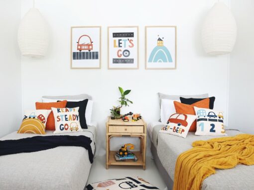 Kids cushions wit cute car designs on them in blues and reds sitting on twin beds with a mustard and navy throw blanket. On the wall are three kids wall prints that match the designs on the cushions