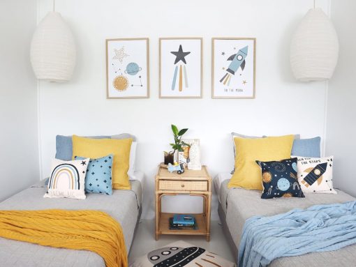 Kids cushions with moon and star prints sitting on two single beds with a mustard throw blanket and a light blue throw. On the wall are three moon and star wall prints that match the cushions