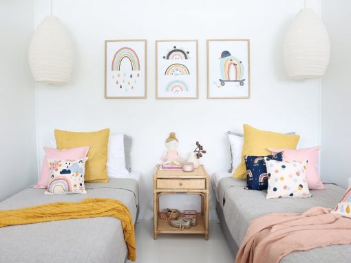 Kids cushions with rainbow prints in blue, pink and navy colours sitting on twin beds with a mustard and pink throw blanket. On the wall are three rainbow kids wall prints that match the cushions