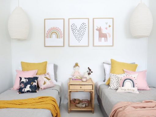 Kids cushions in unicorn pink prints sitting on two single beds with a mustard throw. On the wall are three unicorn prints that match the cushions