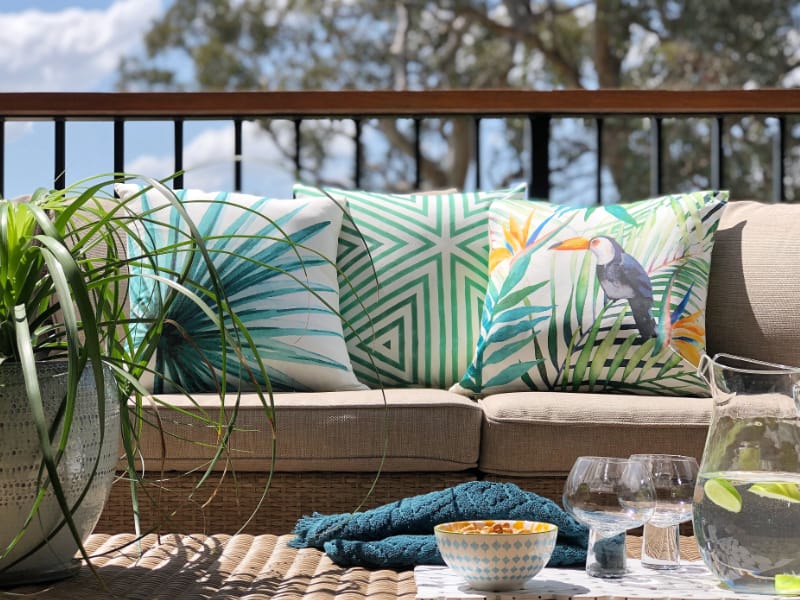 A set of tropical cushions arranged on a lounge in an outdoor scene with trees in the background and food on a table in the foreground.
