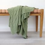 An image of a sage green knitted throw blanket draped over a stool