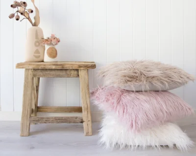 A stack of faux fur fluffy cushion covers in white, pink and light brown sit next to a wooden table.