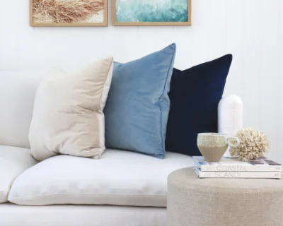 Large cushions in beige, blue and navy sit on a light cream sofa in a coastal styled room.