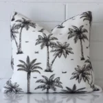 White wall with a palm tree black and white cushion laying against it. It has a distinctive outdoor fabric and has a square shape.