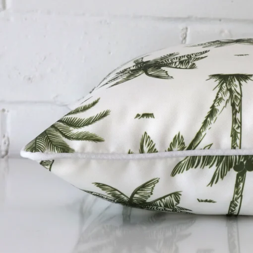 Olive green palm tree cushion cover laying sideways against brick wall. The square size and outdoor material are shown highlighting the seams.