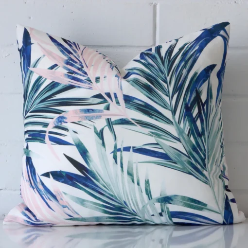 Bold square pink cushion positioned in front of white brickwork. Its palm style pops on the outdoor fabric.