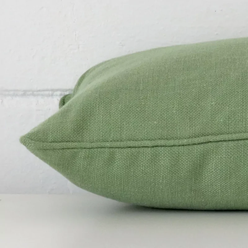 Sage green cushion cover laying sideways against brick wall. The rectangle size and linen material are shown highlighting the seams.