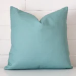 Linen square cushion in an upright position against a white brick wall. It is teal in colour.