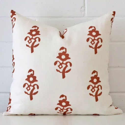 Linen square cushion with a patterned design in an upright position against a white brick wall. It is terracotta in colour.