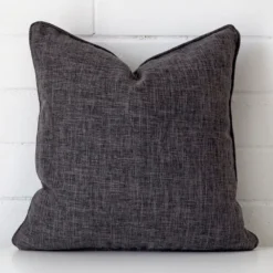 Grey cushion cover in front of a white wall. It has a square size and is made from a linen material.