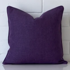 An attractive linen cushion in front of a white brick wall. It has a square shape and is plum in colour.