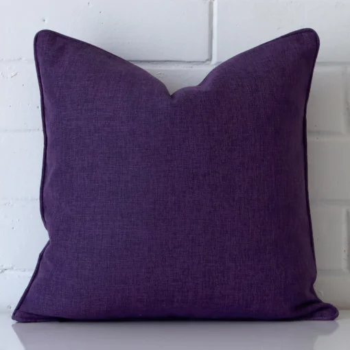 An attractive linen cushion in front of a white brick wall. It has a square shape and is plum in colour.