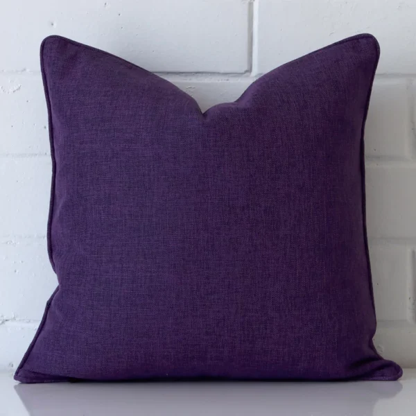 An attractive linen cushion in front of a white brick wall. It has a square shape and is plum in colour.