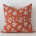 White wall with a floral rust cushion laying against it. It has a distinctive linen fabric and has a square shape.