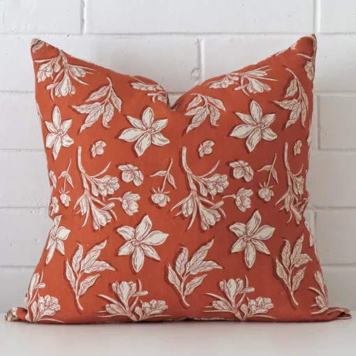 White wall with a floral rust cushion laying against it. It has a distinctive linen fabric and has a square shape.