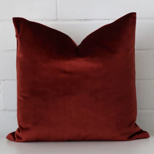 Velvet cushion with a plain design in an upright position against a white brick wall. It is maroon in colour.