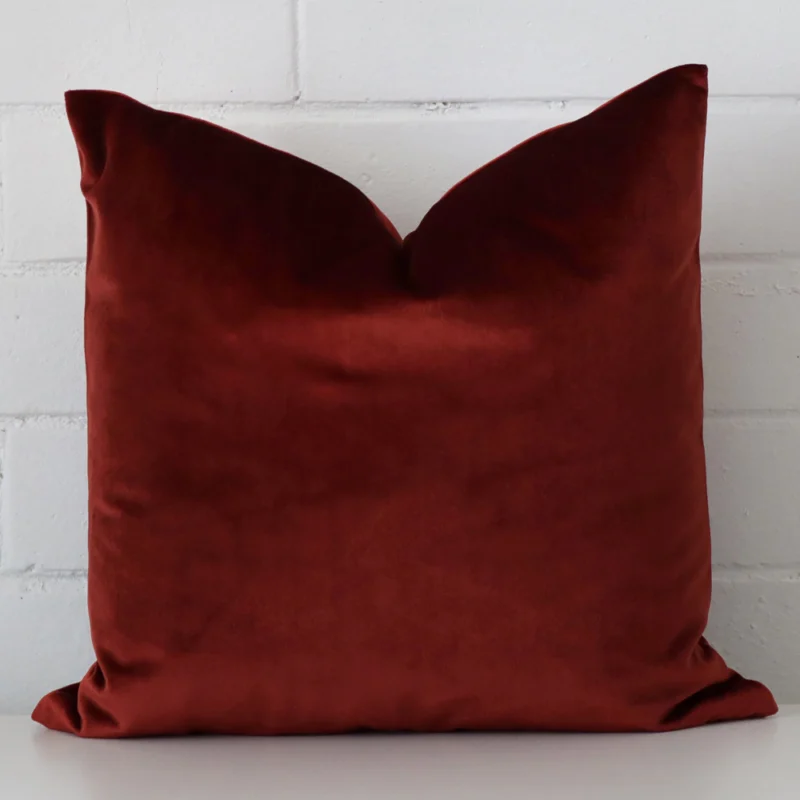 audrey-linen-rust-floral-cushion-cover-large-sc1051-45b Velvet cushion with a plain design in an upright position against a white brick wall. It is maroon in colour.