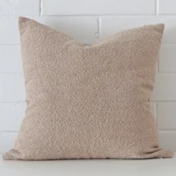 White wall with a beige cushion laying against it. It has a distinctive boucle fabric and has a square shape.