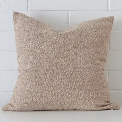 White wall with a beige cushion laying against it. It has a distinctive boucle fabric and has a square shape.