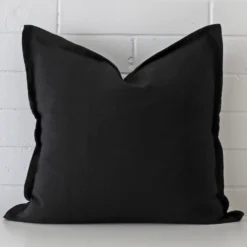 Linen square cushion in an upright position against a white brick wall. It is black in colour.