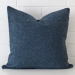 Square cushion cover in blue colour sitting upright in front of a brick wall. It has been made from a quality boucle material.